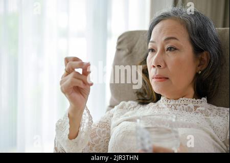 Eine gestresste und depressive asiatische Frau mittleren Alters nimmt in ihrem Wohnzimmer ein Medikament oder eine Pille ein. Konzept der Gesundheitsprobleme. Stockfoto