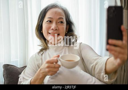 Eine charmante asiatische Frau mittleren Alters, die sich in ihrem Wohnzimmer entspannt, einen Kaffee am Morgen hat und ihr Selfie mit ihrem Smartphone macht. Stockfoto