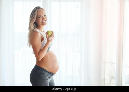 Mein Baby liebt es, wenn ich gut esse. Porträt einer glücklichen Schwangeren, die in Sportkleidung gekleidet ist und zu Hause einen Apfel isst. Stockfoto