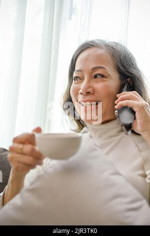 Porträt, Eine glückliche und attraktive asiatische Frau mittleren Alters genießt es, am Telefon mit ihrem Freund zu sprechen, während sie Morgenkaffee in ihrem Wohnzimmer schlürft. Stockfoto
