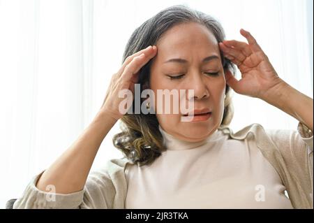 Attraktive asiatisch-gealterte Frau, die an Kopfschmerzen oder Migräne leidet, während sie sich in ihrem komfortablen Wohnzimmer entspannt. Nahaufnahme Stockfoto