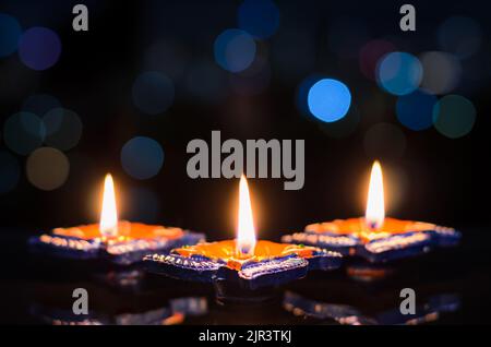 Selektiver Fokus auf die Flamme der ersten Diya-Lampen aus Ton, die auf dunklem Hintergrund mit farbenfrohen Bokeh-Lichtern beleuchtet werden. Diwali Festivalkonzept. Stockfoto