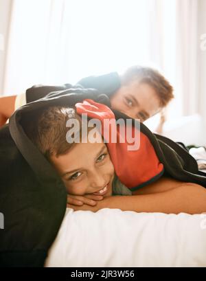 Alle Geschwister haben ihre dummen Momente. Porträt zweier kleiner Brüder, die zu Hause auf einem Bett unter einem Wäschestapel liegen. Stockfoto