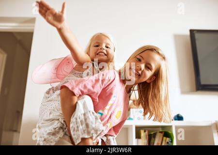 Big sis hilft mir, eine Fee zu werden. Porträt eines kleinen Mädchens, das ihrer Schwester zu Hause eine Huckepack-Fahrt gab. Stockfoto