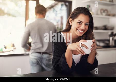 Zeit, um den Tag in Gang zu bringen, aber zuerst Kaffee. Porträt einer jungen Frau, die in der Küche Kaffee getrunken hat, wobei ihr Mann im Hintergrund steht. Stockfoto