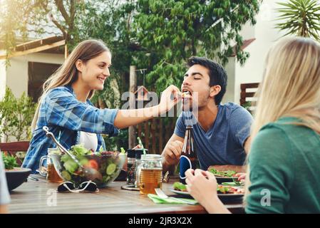 Wie schmeckt mein im Vergleich zu deinem Geschmack? Schoss einen fröhlichen jungen Mann, der von seiner Freundin bei einem Treffen unter Freunden im Freien Pizza gefüttert wurde. Stockfoto