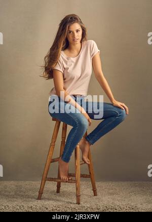 Sie haben, was es braucht. Studioaufnahme einer schönen jungen Frau, die auf einem Hocker vor einem schlichten Hintergrund sitzt. Stockfoto