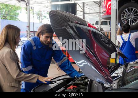 Junger Mann Mechaniker im Gespräch mit dem Kunden für die Untersuchung von Problemen und Reparatur Motor des Autos in der Garage, Fahrzeug der asiatischen Frau mit Problem und Haupt Stockfoto
