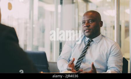Afroamerikanischer Geschäftsmann in formeller Kleidung gestikuliert und erklärt seinen Startup-Plan, um sich während der Mittagszeit in einem glasigen Café zu partnieren Stockfoto