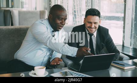 Fröhlicher afroamerikanischer Geschäftsmann in formeller Kleidung, der während der Mittagspause mit seinem kaukasischen Kollegen in einem stilvollen Café über ein Geschäftsprojekt auf seinem Laptop diskutiert Stockfoto