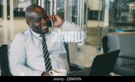 Porträt eines afroamerikanischen Geschäftsmannes in formeller Kleidung, der auf seinem Smartphone spricht und im Internet surft, während er während der Mittagszeit seinen Laptop in einem modernen Café ansieht. Er sieht ehrgeizig und erfolgreich aus. Stockfoto