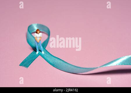 Miniatur Menschen Figur Spielzeug Fotografie. Konzept des Ovarian Cancer Awareness Day. Mädchen, Teenager, Frau, die über grün türkisfarbenem blauem blauem Band steht. Bild Stockfoto