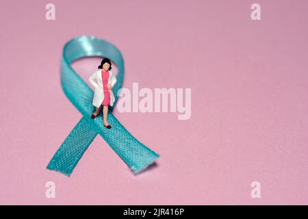 Miniatur Menschen Figur Spielzeug Fotografie. Konzept des Ovarian Cancer Awareness Day. Mädchen Frau Arzt steht über grün türkis blaues blaues blaues Band. Bild-ph Stockfoto