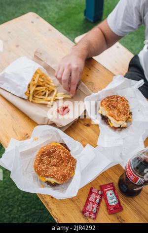 Mann am Tisch im Freien mit Pommes zum Teilen, Heinz Ketchup auf braunem Papierbeutel und zwei Cheeseburgern mit Sesambrötchen und einer Flasche Cola Stockfoto