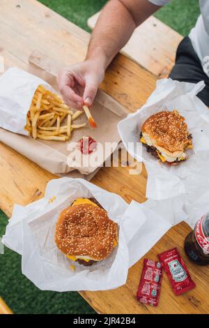 Mann am Tisch im Freien mit Pommes zum Teilen, Heinz Ketchup auf braunem Papierbeutel und zwei Cheeseburgern mit Sesambrötchen und einer Flasche Cola Stockfoto