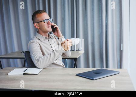 Innenaufnahme des fröhlichen bärtigen männlichen Freiberuflers Arbeit in fernen, verwaltet Finanzen über Smartphone, sitzt am Holztisch, arbeitet mit Dokumentation, hat Stockfoto