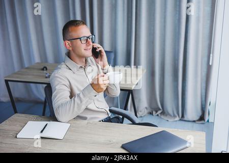 Innenaufnahme des fröhlichen bärtigen männlichen Freiberuflers Arbeit in fernen, verwaltet Finanzen über Smartphone, sitzt am Holztisch, arbeitet mit Dokumentation, hat Stockfoto