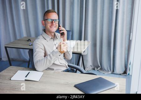 Innenaufnahme des fröhlichen bärtigen männlichen Freiberuflers Arbeit in fernen, verwaltet Finanzen über Smartphone, sitzt am Holztisch, arbeitet mit Dokumentation, hat Stockfoto