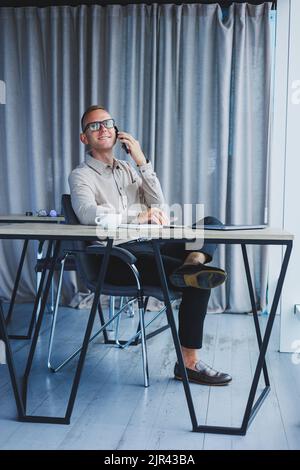 Innenaufnahme des fröhlichen bärtigen männlichen Freiberuflers Arbeit in fernen, verwaltet Finanzen über Smartphone, sitzt am Holztisch, arbeitet mit Dokumentation, hat Stockfoto