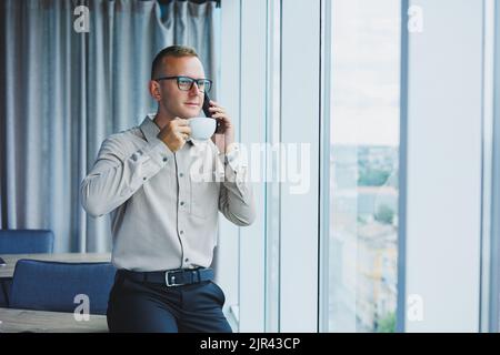 Innenaufnahme des fröhlichen bärtigen männlichen Freiberuflers Arbeit in fernen, verwaltet Finanzen über Smartphone, sitzt am Holztisch, arbeitet mit Dokumentation, hat Stockfoto