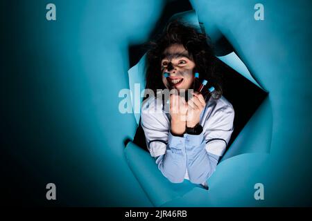Verrückter Wissenschaftler mit unordentlicher Frisur und gefüllten Glasröhrchen, die auf blauem Hintergrund schrecklich vor der Kamera grinsen. Dumme, wahnsinnige Chemikerin mit einem Tablett mit Reagenzgläsern, die mit giftigen Verbindungen gefüllt sind Stockfoto
