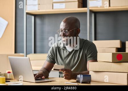 Afrikanischer Lagerarbeiter, der online mit der Eingabe der Kreditkarte auf dem Laptop am Tisch bezahlt Stockfoto