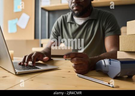 Nahaufnahme eines afrikanischen Mannes, der am Tisch sitzt und mit einem Laptop online mit Kreditkarte bezahlt Stockfoto