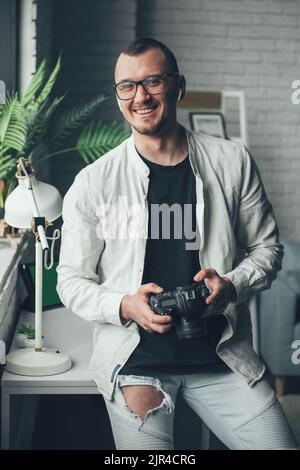 Fröhlicher Fotograf, der die Kamera lächelnd in seinem Büro vor der Kamera hält. Freiberufliche kreative Arbeit Online-Marketing. Gründung des Inhabers von Kleinunternehmen. Stockfoto