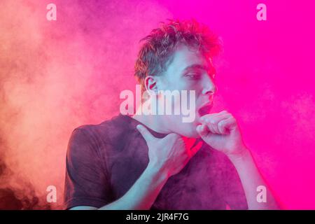 Ersticken Sie im Rauch. Ängstlicher junger Mann mit gestressten Emotionen isoliert über rosa Hintergrund mit Wolken aus Rauch. Konzept des Gesichtsausdrucks Stockfoto