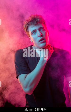 Ersticken Sie im Rauch. Ängstlicher junger Mann mit gestressten Emotionen isoliert über rosa Hintergrund mit Wolken aus Rauch. Konzept des Gesichtsausdrucks Stockfoto