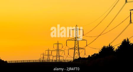 Strommasten bei Sonnenuntergang, Silhouetten gegen Orange Sky Stockfoto