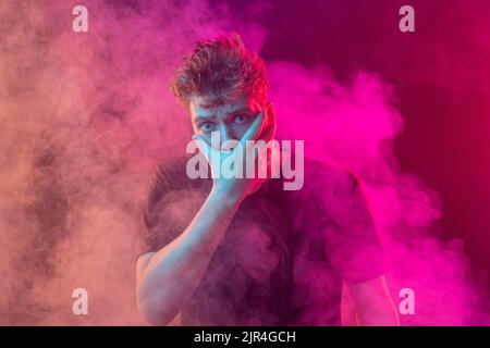 Ersticken Sie im Rauch. Ängstlicher junger Mann mit gestressten Emotionen isoliert über rosa Hintergrund mit Wolken aus Rauch. Konzept des Gesichtsausdrucks Stockfoto