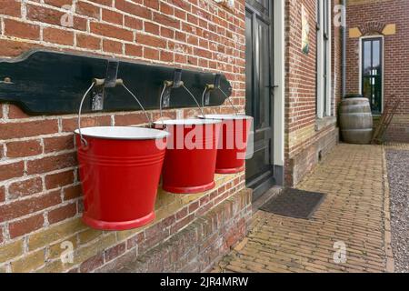 Drei rote Metall-Feuereimer, die in einer Reihe an einer Backsteinmauer an einem alten Bahnhof hängen. Stockfoto
