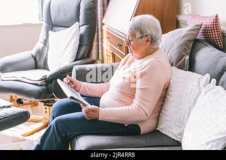 Alte ältere Frau, 80s Erwachsene, Großmutter, die Kreuzworträtsel in ihrem Wohnzimmer macht, das auf dem Sofa sitzt. Ruhestandhobby für Trainingsgedächtnis. Alzhe Stockfoto