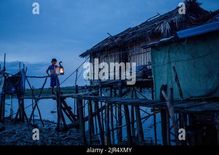 Kalabogi, Bangladesch. 18. August 2022. Ein Junge sah, wie er im Dorf Kalabogi in Khulna eine Bambusbrücke überquerte, während er eine Laterne an der Hand hielt. Vor nicht allzu langer Zeit war Kalabogi, ein Küstendorf in Bangladesch, voller anbauflächiger Flächen, bis der steigende Meeresspiegel das Gebiet bis zur Bucht von Bengalen verschlang. Seit Ende 1990s ereignissen sich im Dorf häufige Wirbelstürme und Überschwemmungen. Im Jahr 2009 zerstörte ein großer Wirbelsturm namens Aila die 1.400 Kilometer Böschungen, 8.800 Kilometer Straßen und etwa 3 50.000 Hektar Ackerland des Landes. Mehrere hundert Menschen seien dabei getötet worden Stockfoto