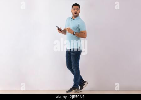 Porträt eines überraschten jungen Mannes, der mit Smartphone und Kaffee vor weißem Hintergrund steht Stockfoto
