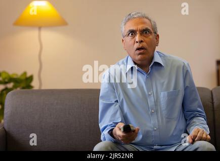 Schockierter älterer Mann, der im Wohnzimmer auf dem Sofa saß und Fernsehen schaute. Stockfoto