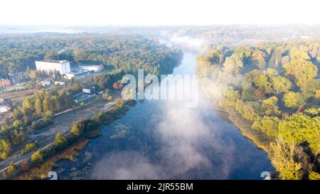 Luftdrohnen-Ansicht, die an einem sonnigen frühen Herbstmorgen über den Fluss fliegen, mit ruhiger reflektierender Wasseroberfläche und weißem Nebelnebel und Waldbäumen am Flussufer. Schöner natürlicher Hintergrund. Stockfoto