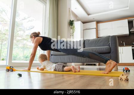 Seitenansicht einer jungen, gesunden Mutter, die beim Training stand und in Plankenposition auf einer gelben Yogamatte neben einem großen Fenster zu Hause stand, wobei ein süßer Junge in ihrer Nähe lag. Glückliche Mutterschaft und postnataler Sport Stockfoto