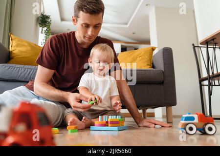 Vater unterstützt seinen kleinen Jungen beim Sortierer, der auf dem Boden im Wohnzimmer mit Sofa auf dem Hintergrund sitzt, umgeben von Autospielzeugen, und wartet darauf, dass seine Mutter von der Arbeit nach Hause kommt. Schulspielzeug, Kinderentwicklung Stockfoto