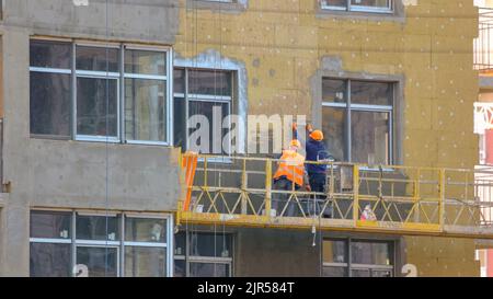 Arbeiter isolieren die Wand eines Gebäudes. Baustelle mit arbeitenden Männern auf dem Aufzug. Paneele. Aufgehängte Aufzugsvorrichtung. Stockfoto