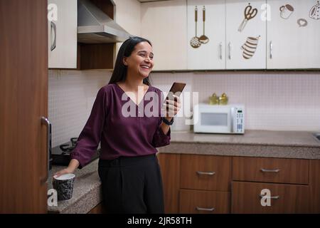 Lächelnde Frau mit einer Tasse Kaffee und Smartphone in der Küche Stockfoto