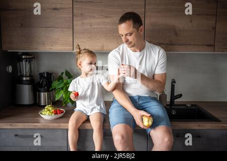 Nettes kleines Mädchen und ihr hübscher Vater essen Obst in der modernen Küche. Gesunde Ernährung. Stockfoto