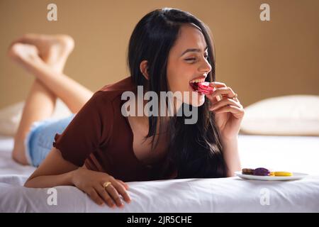 Glückliche junge Frau, die Makronen isst, während sie sich auf das Bett legt Stockfoto