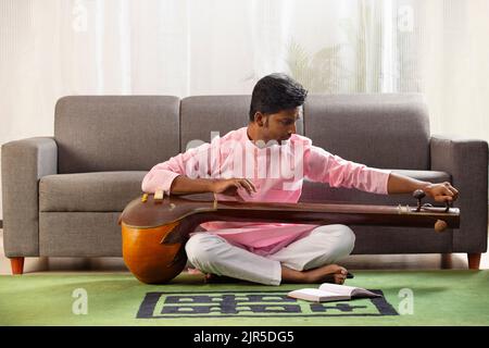 Junger Musiker stimmt seine Tanpura, während er im Wohnzimmer sitzt Stockfoto