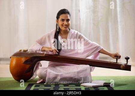 Junge Musikerin stimmt ihr Tanpura, während sie im Wohnzimmer sitzt Stockfoto