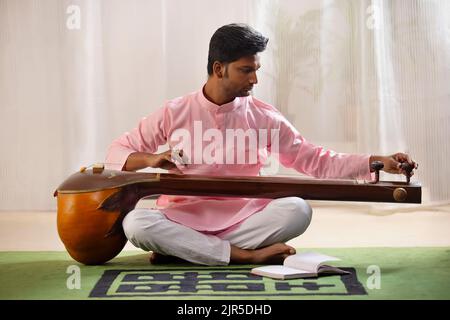 Junger Musiker stimmt seine Tanpura, während er im Wohnzimmer sitzt Stockfoto