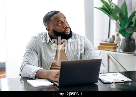 Nachdenklich verärgert afroamerikanischer Geschäftsmann, ceo, Büroangestellter, sitzt im modernen Büro an einem Tisch, an seinem Arbeitsplatz, schaut nachdenklich zur Seite, denkt über ein neues Projekt nach, träumt von Ruhe, Urlaub Stockfoto