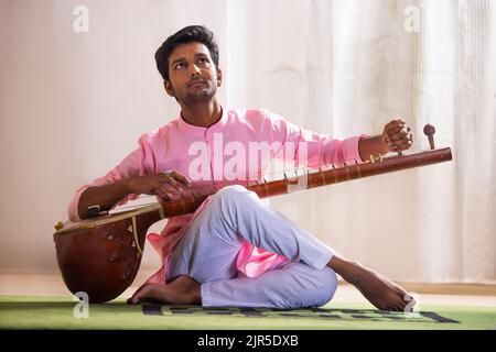 Junger Musiker, der seine Sitar stimmt, während er im Wohnzimmer sitzt Stockfoto