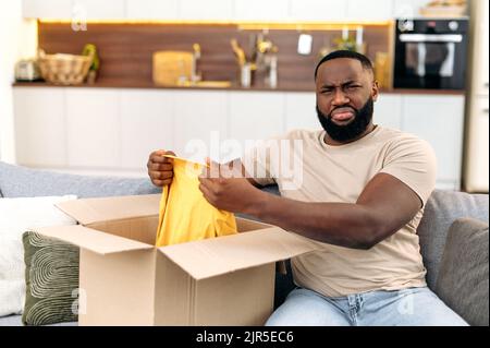 Frustrierter afroamerikanischer Mann in legerer Kleidung, sitzt auf dem Sofa im Wohnzimmer, packt sein Paket aus, nimmt Kleidung aus dem Karton, schaut enttäuscht und unzufrieden auf die Kamera Stockfoto